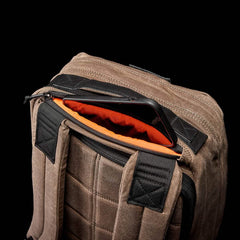 A close-up shot features a brown GR1 USA - Heritage by GORUCK, showcasing its black straps and partially open design that reveals a tablet inside. The interior lining is vibrant orange, providing contrast to the rugged exterior made from waxed duck canvas. The background is solid black.