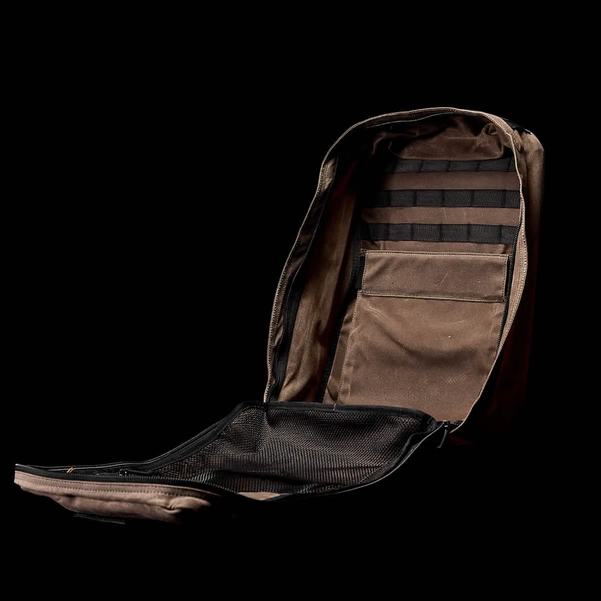 The GR1 USA - Heritage by GORUCK is an open tactical backpack crafted from brown waxed duck canvas, set against a black background. It includes multiple compartments, such as a zippered mesh pocket and elastic straps inside, highlighting its organized and utilitarian design.