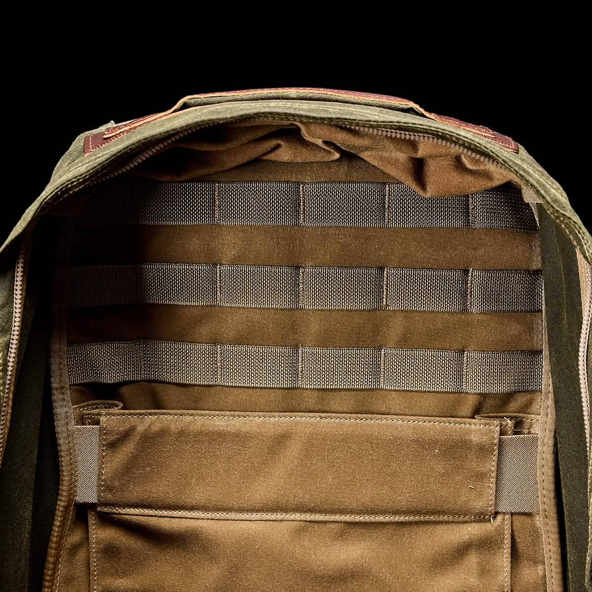 The image showcases a GORUCK GR1 USA - Heritage Backpack with an open view of its interior. The inside is brown, featuring horizontal straps and a large pocket for organizing belongings. The exterior is crafted from olive green fabric, echoing a retro canvas rucksack style with visible zippers.