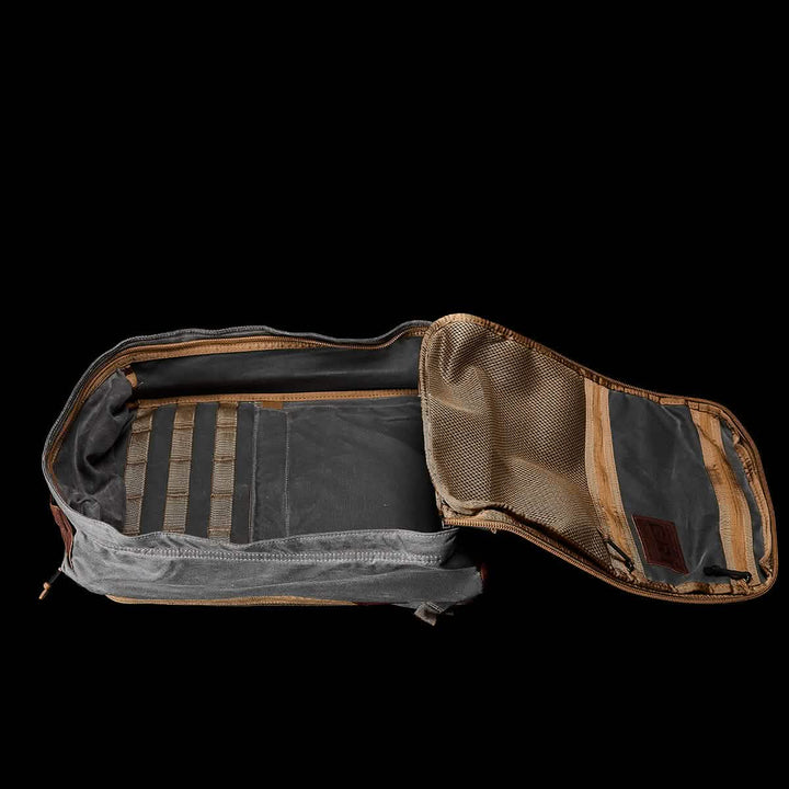 Display the GORUCK GR1 USA - Heritage backpack in black and brown against a black backdrop, highlighting its waxed canvas exterior with a variety of interior pockets and compartments. The open flap reveals mesh pockets and zippers, tailored for optimal organization.
