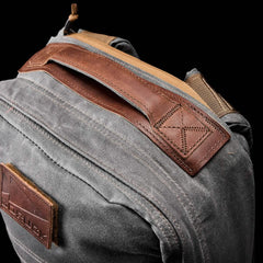 Close-up of a rugged gray GR1 USA - Heritage rucksack from GORUCK featuring a brown leather handle and tan accents. The waxed canvas texture and precise stitching enhance its durability. The black background emphasizes the backpack’s detailed design.