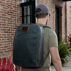A man wearing a gray cap and sunglasses carries the GR1 USA - Heritage rucksack by GORUCK, crafted from waxed canvas and featuring a leather patch. He stands outdoors near a brick building with black-framed windows and a red Adirondack chair.