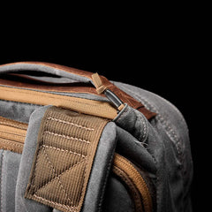 Close-up of the GORUCK GR1 USA - Heritage, a gray waxed canvas rucksack adorned with brown leather accents and a durable brown strap. The detailed stitching and zipper stand out against a black background, capturing the rugged elegance of its design.