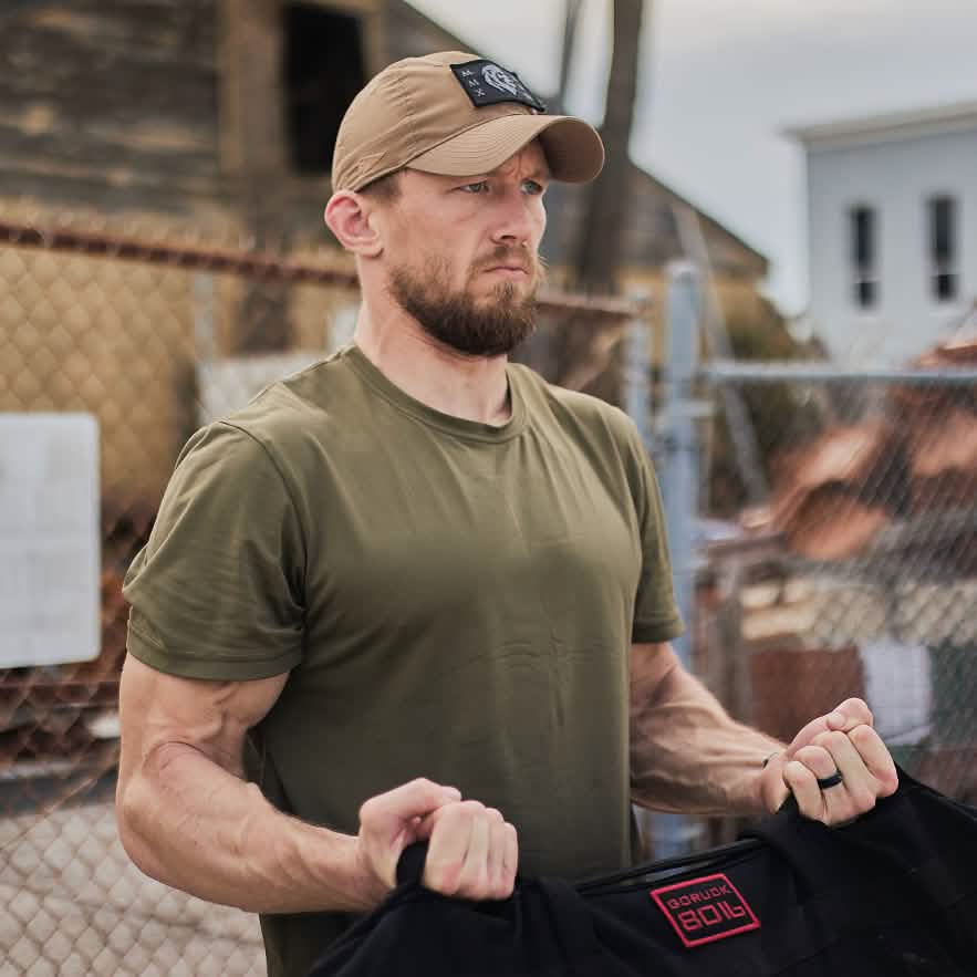 A man in an olive green shirt and beige cap holds a black product emblazoned with red letters "GORUCK" while outdoors, ready for high-speed dynamic movements. A chain-link fence and rustic building serve as the backdrop to his focused expression, embodying the intensity of the High Speed, Low Drag Training Program by GORUCK.