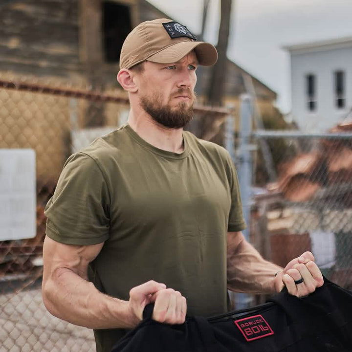 A man in an olive green shirt and beige cap holds a black product emblazoned with red letters "GORUCK" while outdoors, ready for high-speed dynamic movements. A chain-link fence and rustic building serve as the backdrop to his focused expression, embodying the intensity of the High Speed, Low Drag Training Program by GORUCK.