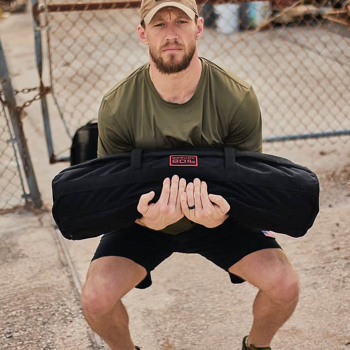 A man wearing a green shirt and cap performs dynamic movements while squatting with a heavy black sandbag outdoors near a chain-link fence, in what looks like an urban or industrial environment. This setting is ideal for incorporating the High Speed, Low Drag Training Program by GORUCK into his fitness routine.
