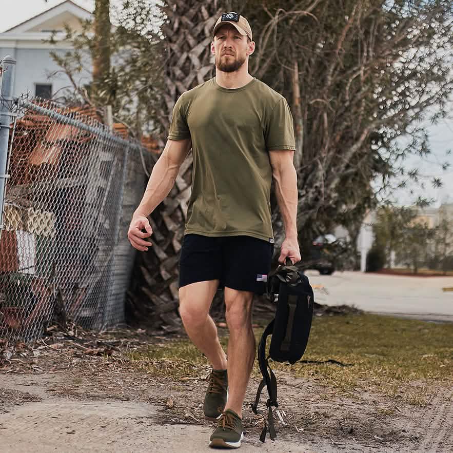 A man wearing a khaki t-shirt, black shorts, and a baseball cap swiftly navigates the dirt path with dynamic movements. He carries a GORUCK backpack in one hand, showcasing the High Speed, Low Drag Training Program as trees and a chain-link fence frame the background.