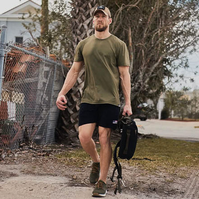 A man wearing a khaki t-shirt, black shorts, and a baseball cap swiftly navigates the dirt path with dynamic movements. He carries a GORUCK backpack in one hand, showcasing the High Speed, Low Drag Training Program as trees and a chain-link fence frame the background.