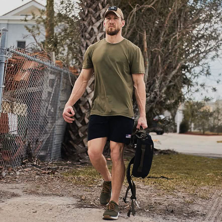 A man wearing a khaki t-shirt, black shorts, and a baseball cap swiftly navigates the dirt path with dynamic movements. He carries a GORUCK backpack in one hand, showcasing the High Speed, Low Drag Training Program as trees and a chain-link fence frame the background.