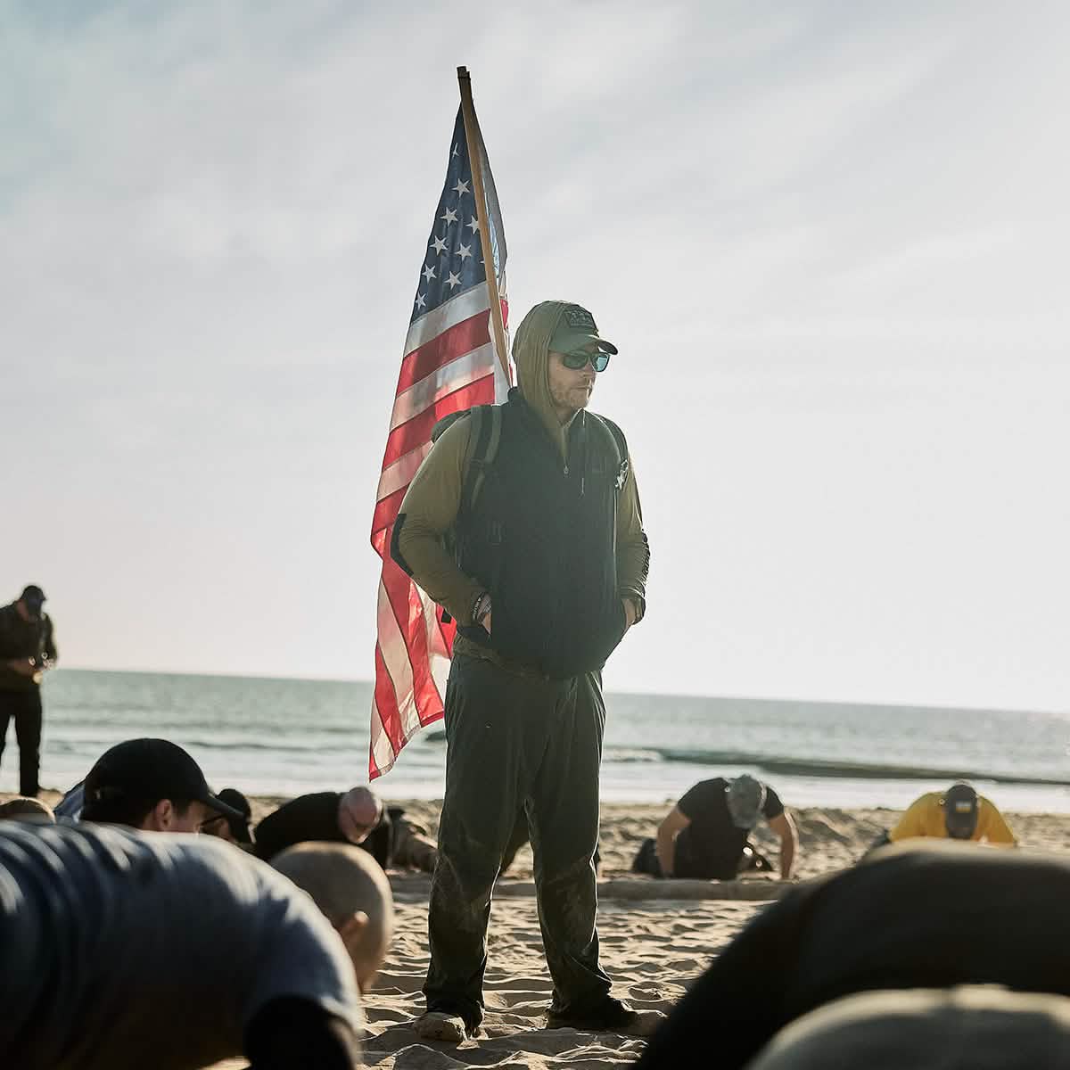 A person stands on a sandy beach holding an American flag, wearing a hooded jacket and sunglasses. Several others in GORUCK Men's Commando Pullovers made from Merino wool are doing exercises on the sand, with the ocean in the background under a partly cloudy sky.