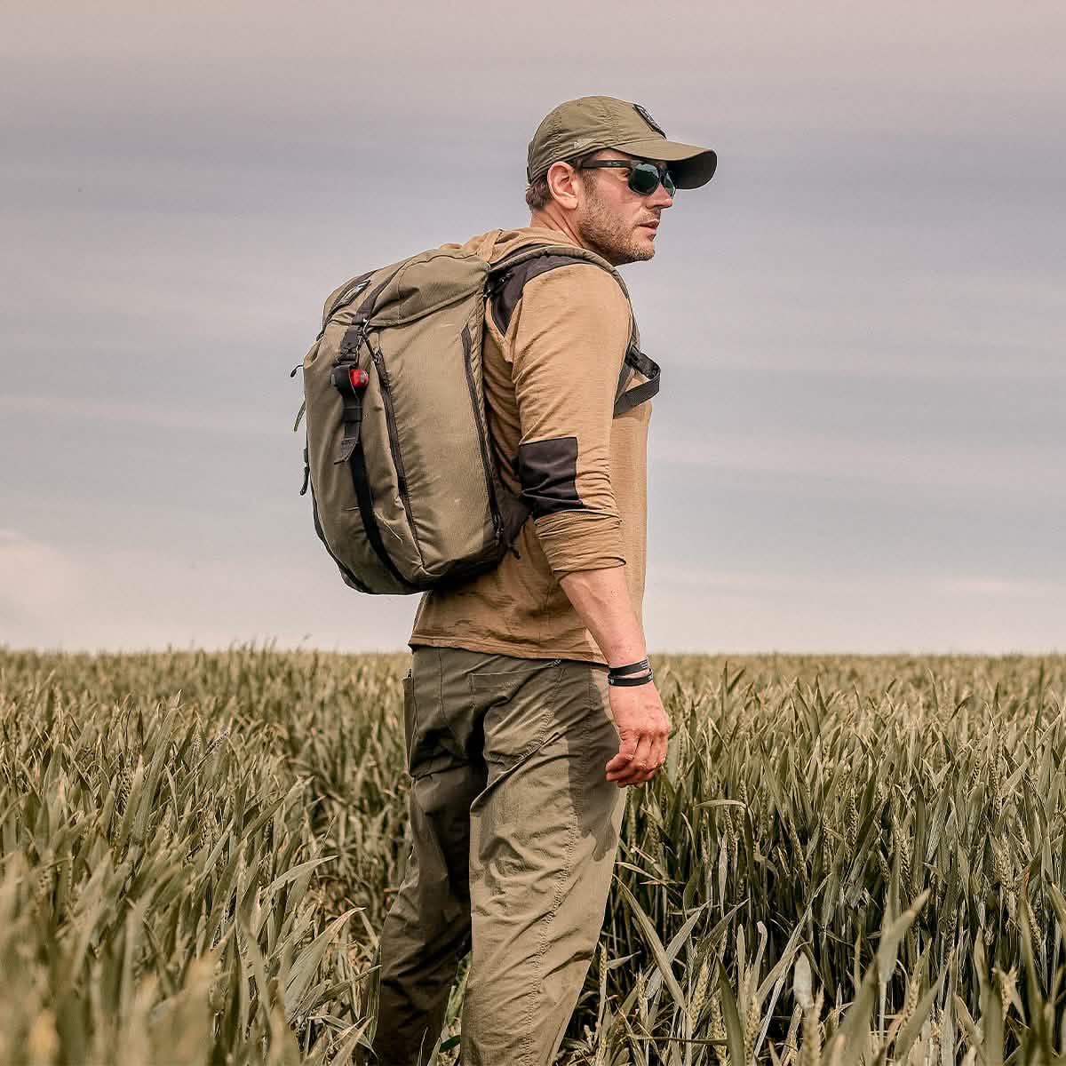 A man wearing a hat, sunglasses, and a backpack stands in a field with tall grass, gazing into the distance. The sky is overcast, and he is dressed in outdoor attire featuring the GORUCK Men’s Commando Pullover made from Merino Wool.