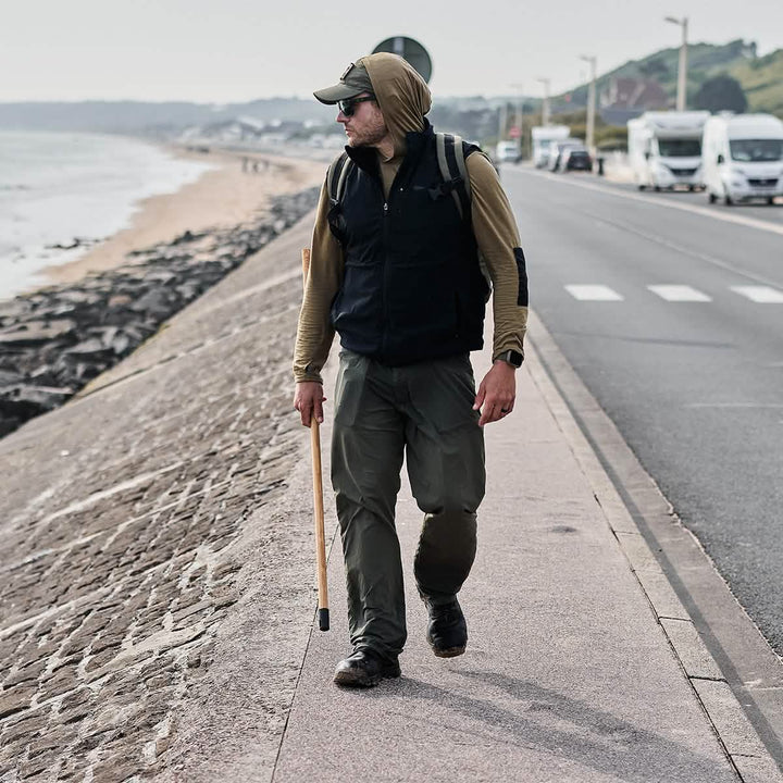 A man dressed in outdoor attire, featuring a GORUCK Men’s Commando Pullover made from merino wool and a matching hat, strolls along a seaside promenade with a walking stick in hand. Parked vans line the road while waves gently crash on the sandy beach in the background. The atmosphere is overcast yet serene, evoking memories of British Commando Sweater days.
