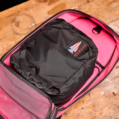 An open Bullet Ruck Laptop - 14L from GORUCK sits on a wooden table, revealing a black packing cube decorated with an American flag patch and the word GORUCK. The bag's interior features bright pink lining and includes a bombproof compartment for secure laptop storage.