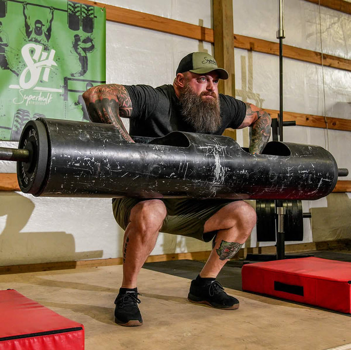A bearded man in a cap, wearing GORUCK Men's Ballistic Trainers - HULK, lifts a large, heavy barbell with both hands in a gym setting. He is squatting and appears focused. The gym features a white wall with a green poster and wooden beams, with red mats placed nearby.