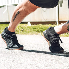 Close-up of a person wearing black rucking shoes and black ankle socks outdoors on pavement, showing calf tattoos