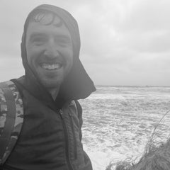 Smiling man in an Indestructible Challenge Windbreaker stands by a rough, windy beach on an overcast day.