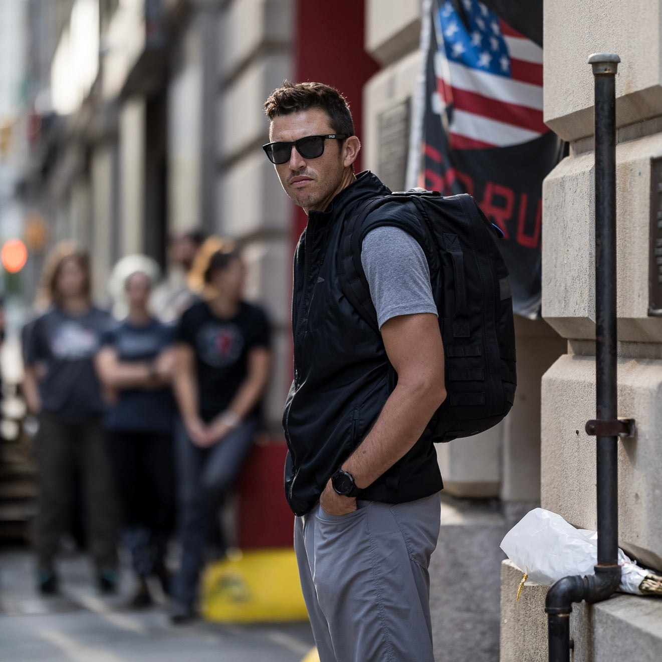 Man in sunglasses and a Men's Vest of Power - ToughDry® + Fleece stands on a city sidewalk with a backpack; others are blurred in the background.