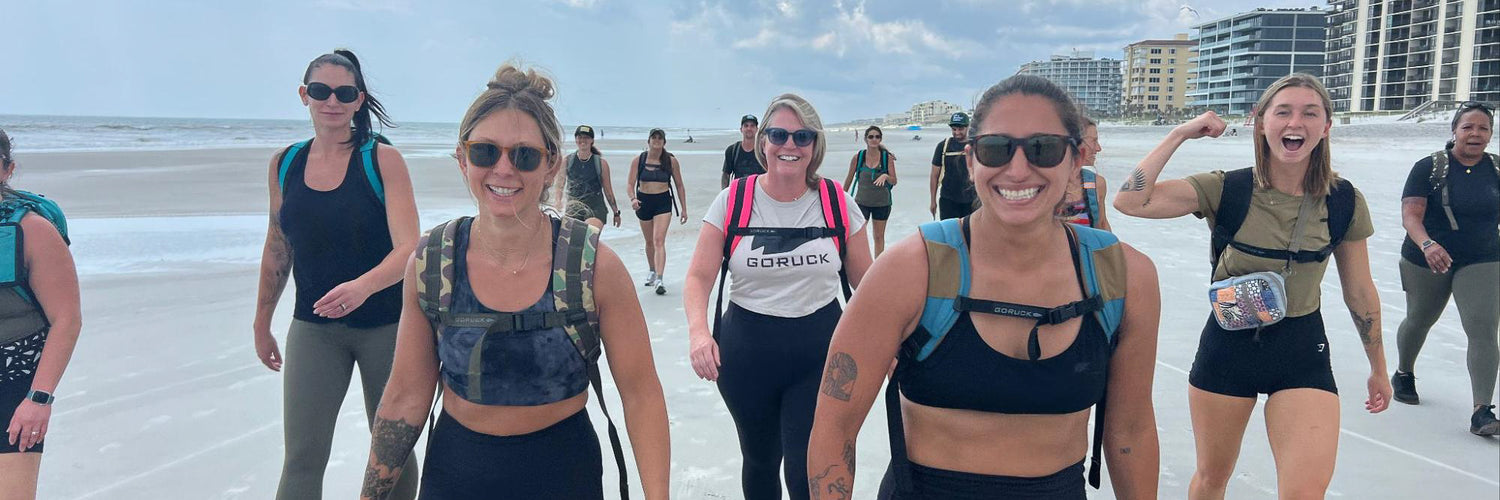 A group of women wearing athletic gear walk and smile together on a sandy beach.
