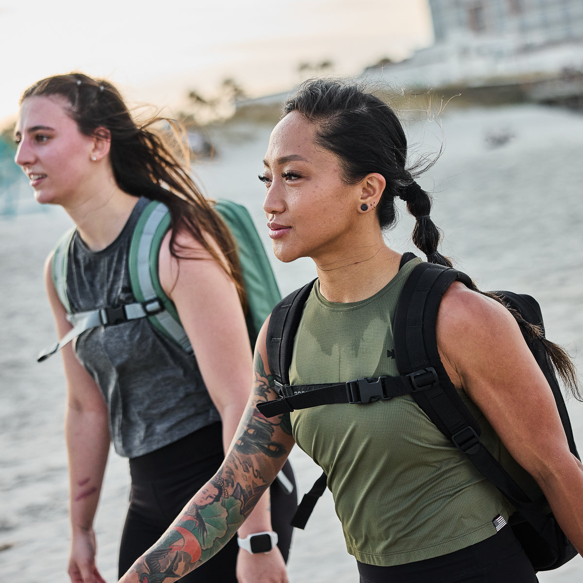 Two women with backpacks walk on a sandy beach, one with a tattooed arm, both wearing athletic clothing.