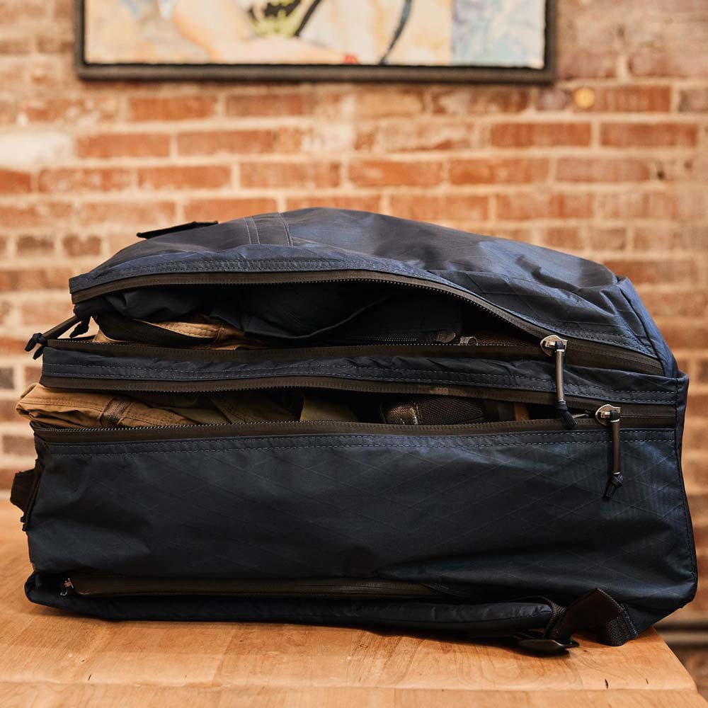 A navy duffel bag sits open and packed with clothes on a wooden table against a brick wall.