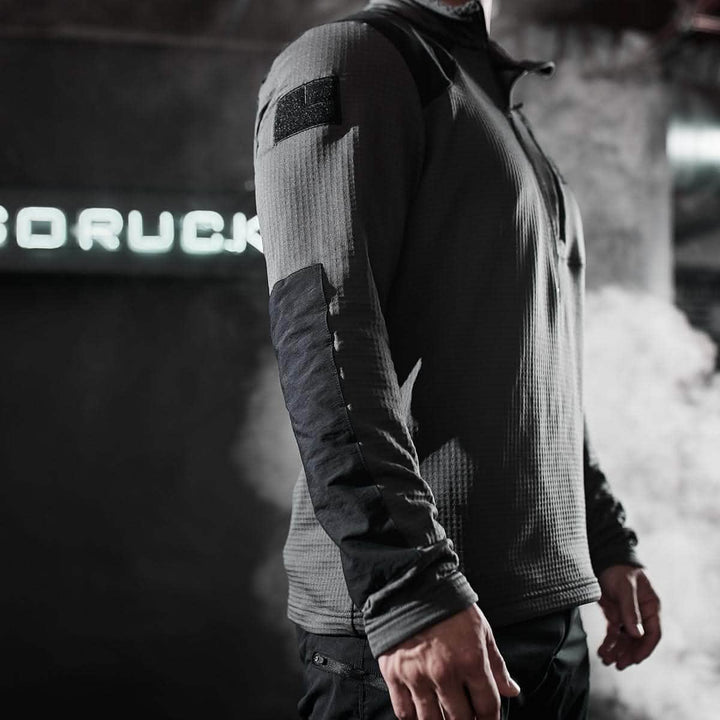 A person wearing the Men’s Half Zip - Polartec Grid Fleece by GORUCK stands in an industrial setting. Steam drifts in the background, partially obscuring a neon sign. The focus is on the jacket's texture and design, featuring a sleek fleece-lined interior for warmth.