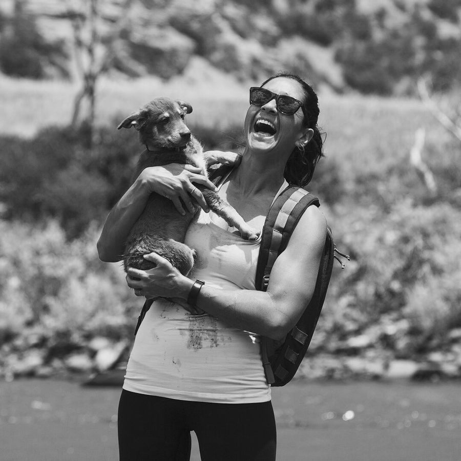 A woman in sunglasses laughs while holding a dog outdoors, wearing a tank top and life vest.