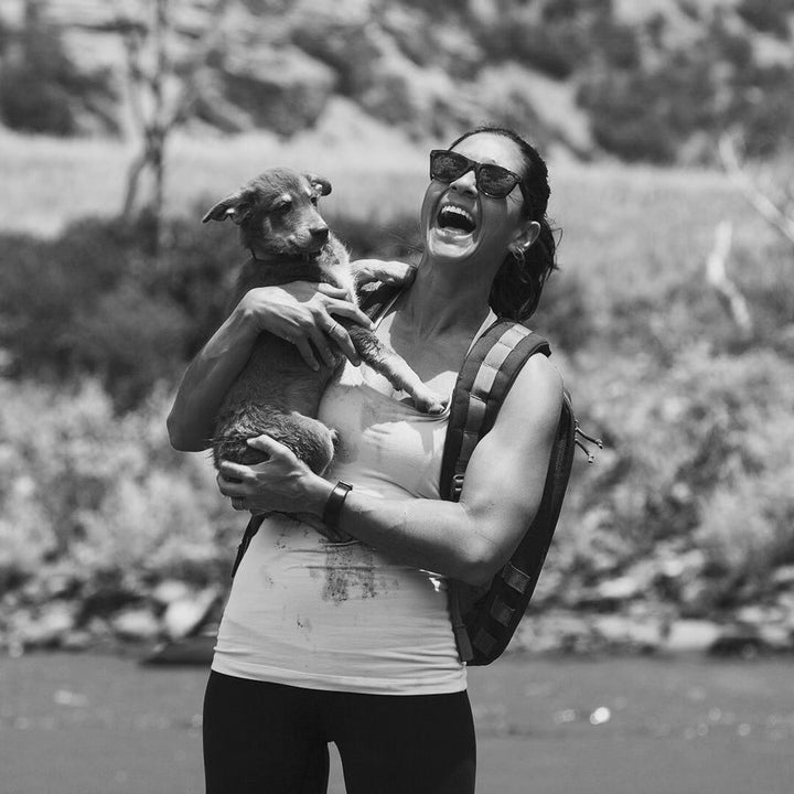 A woman in sunglasses laughs while holding a dog outdoors, wearing a tank top and life vest.