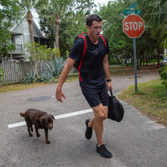 Wearing Men’s Training Shorts - ToughStretch with a built-in liner, a man carries a bag as he walks with a brown dog across a street near a stop sign and palm trees.