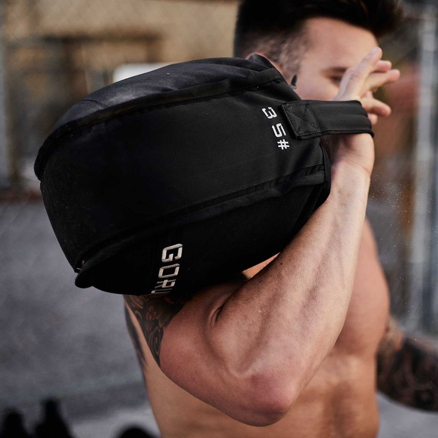 Shirtless person lifting a black 35-pound sandbag over their shoulder outdoors.
