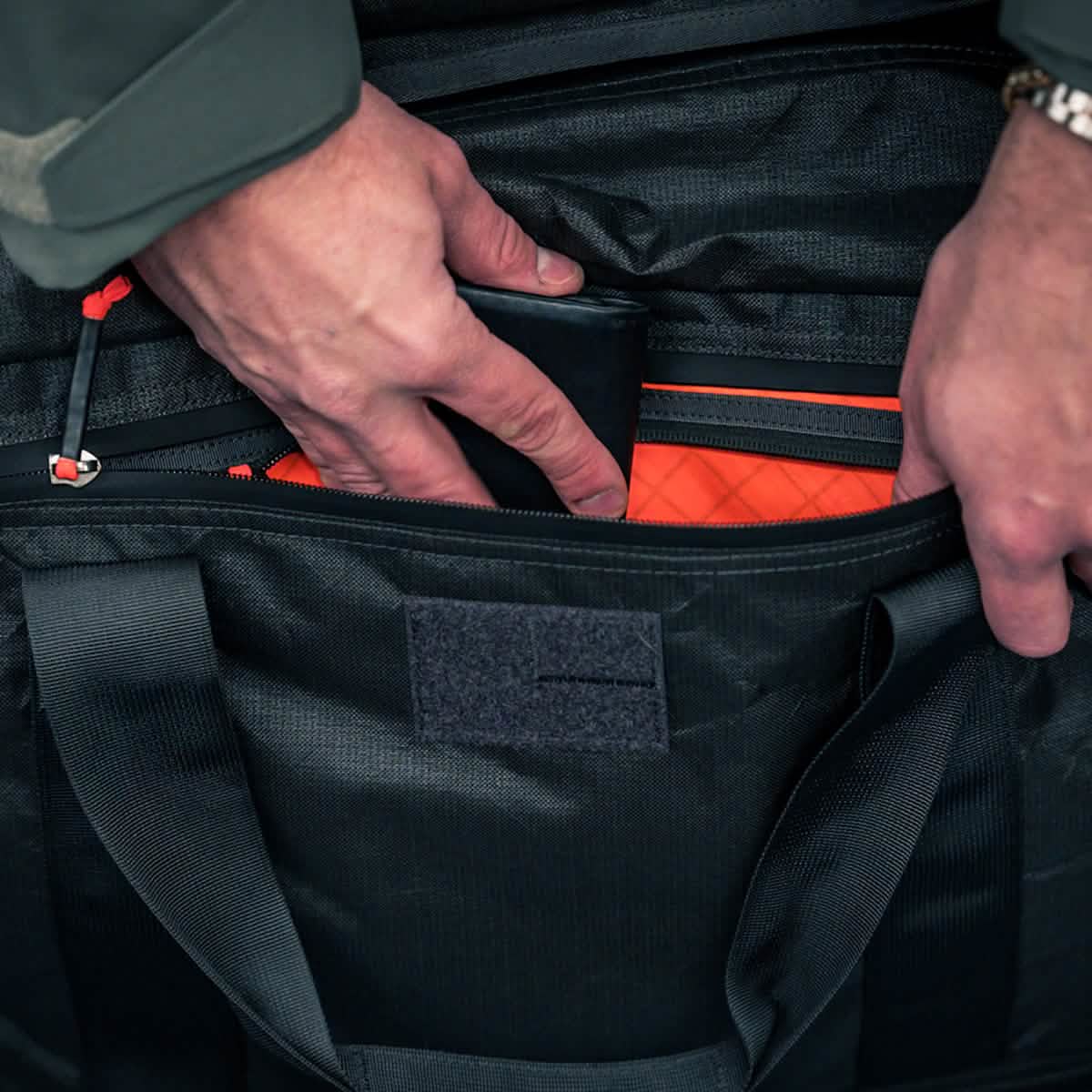 A person places a black wallet into the orange-lined pocket of the Kit Bag x Carryology - Ultra Blaze, which features a shoe compartment.