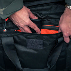 A person places a black wallet into the orange-lined pocket of the Kit Bag x Carryology - Ultra Blaze, which features a shoe compartment.