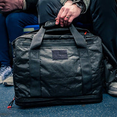 A person holding the Kit Bag x Carryology - Ultra Blaze, a sleek black kit bag with a shoe compartment, while sitting on public transportation.