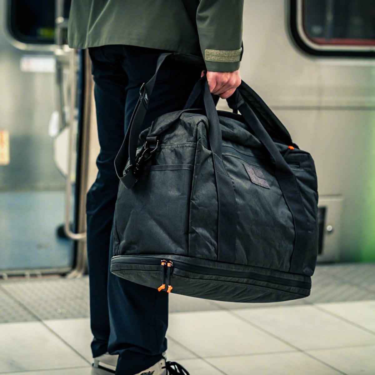Person holding a large black Kit Bag x Carryology - Ultra Blaze duffel made of UHMWPE fabric on a train station platform, with a train in the background.