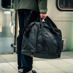 Person holding a large black Kit Bag x Carryology - Ultra Blaze duffel made of UHMWPE fabric on a train station platform, with a train in the background.