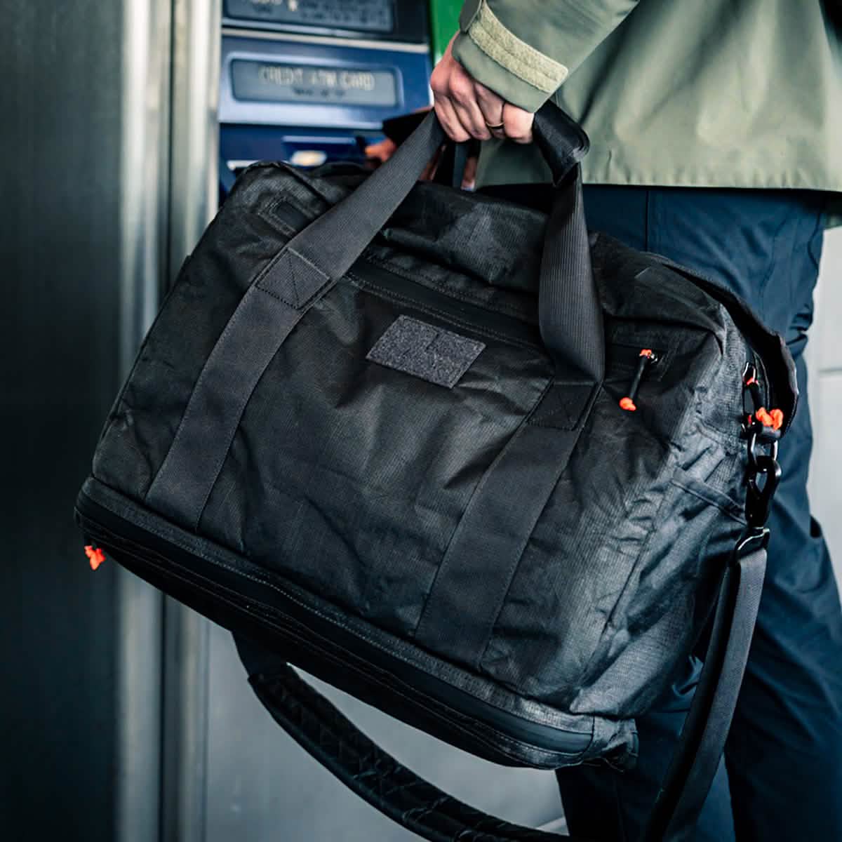 A person stands near an ATM holding the Kit Bag x Carryology - Ultra Blaze, a large black kit bag with a shoe compartment, handles, and a shoulder strap.