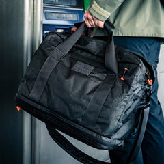 A person stands near an ATM holding the Kit Bag x Carryology - Ultra Blaze, a large black kit bag with a shoe compartment, handles, and a shoulder strap.