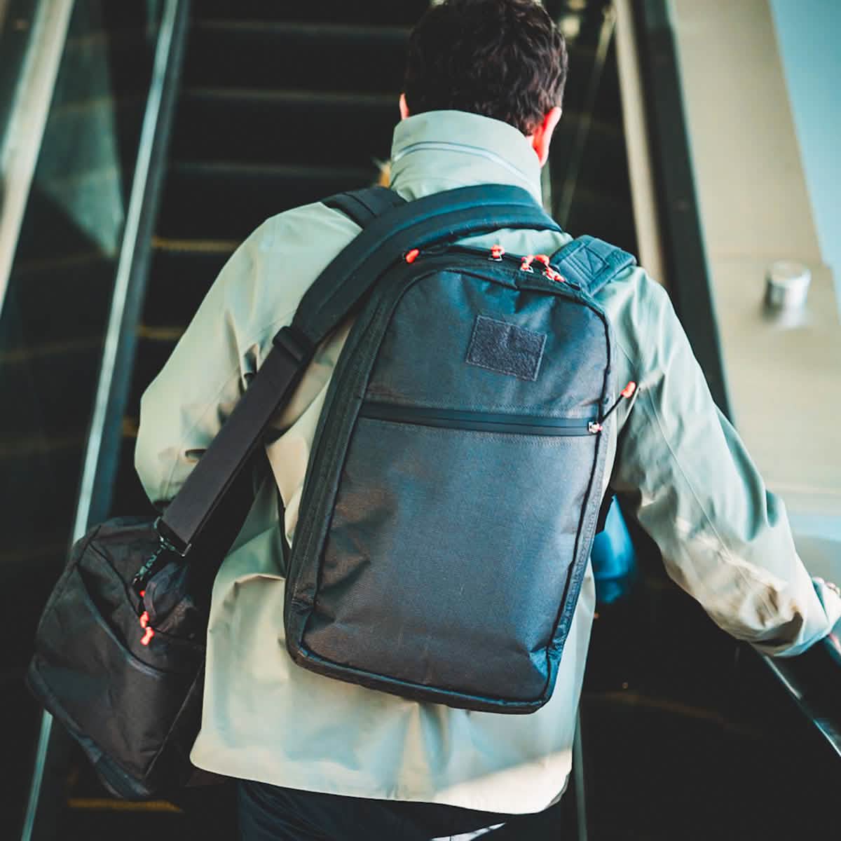 A person with a backpack and the Kit Bag x Carryology - Ultra Blaze, featuring a shoe compartment, rides up an escalator, seen from behind.