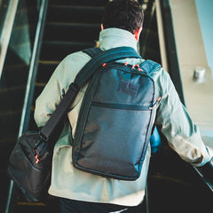 A person with a backpack and the Kit Bag x Carryology - Ultra Blaze, featuring a shoe compartment, rides up an escalator, seen from behind.