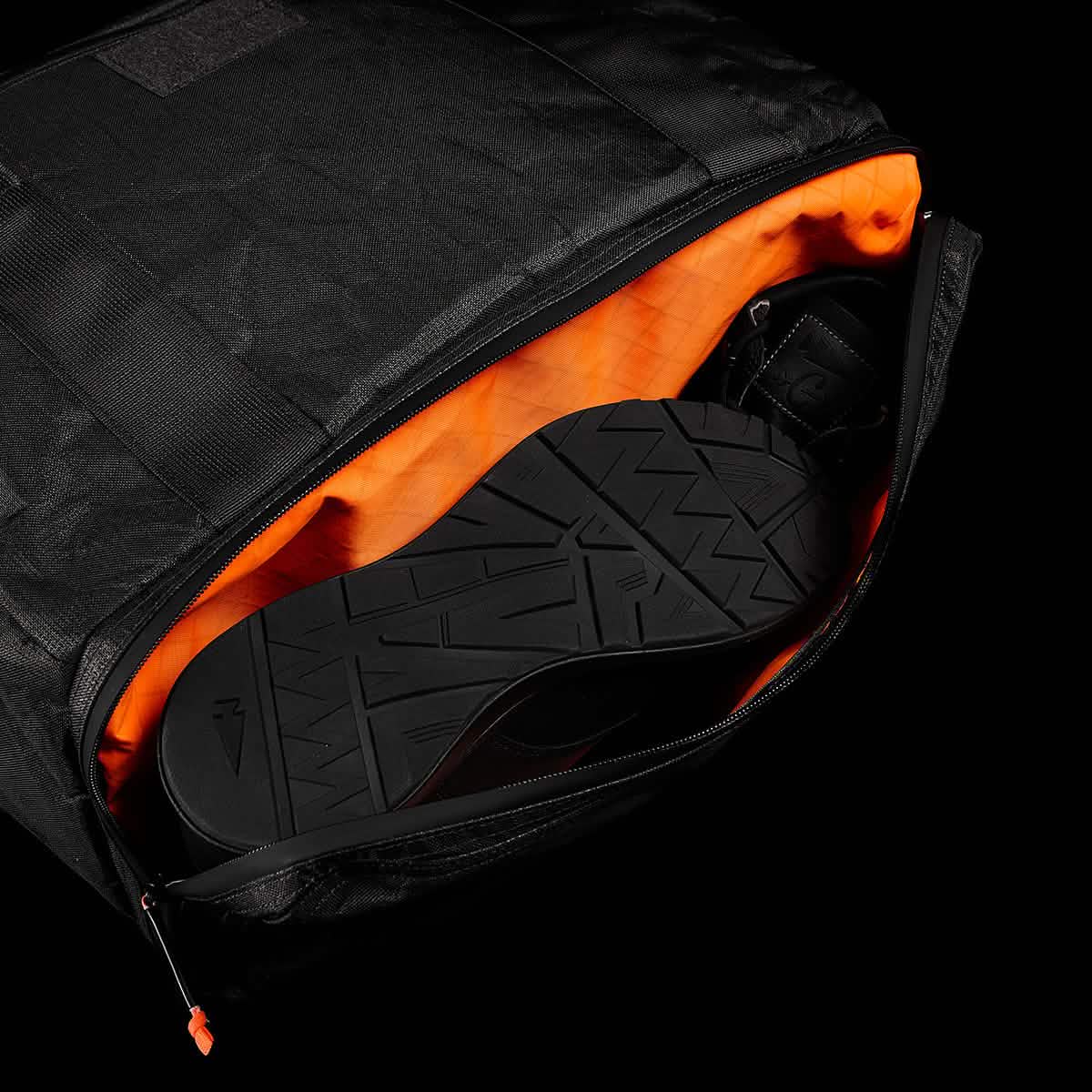 The Kit Bag x Carryology - Ultra Blaze is a sleek black backpack with an orange interior, unzipped to show black shoes inside—a stylish bag featuring a dedicated shoe compartment for organized travel.
