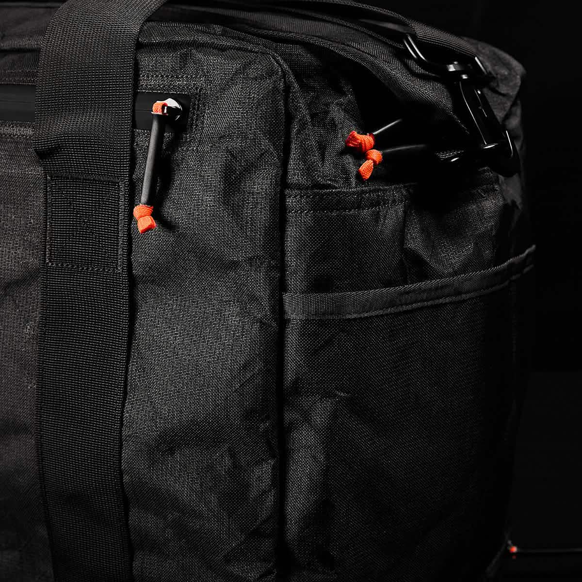 Close-up of the Kit Bag x Carryology - Ultra Blaze duffel, crafted from UHMWPE fabric, with black zippers and orange zipper pulls, set against a dark background.