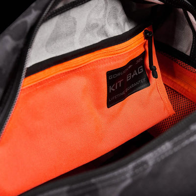 Close-up of an open bag displaying an orange zippered pocket inside. A label on the pocket reads GORUCK Kit Bag: Lifetime Guarantee. The interior of the bag is gray with black accents, and the unzipped zipper reveals a vibrant orange inner pocket, reminiscent of Special Forces Flight Bags.