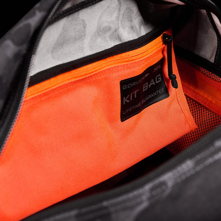 Close-up of an open bag displaying an orange zippered pocket inside. A label on the pocket reads GORUCK Kit Bag: Lifetime Guarantee. The interior of the bag is gray with black accents, and the unzipped zipper reveals a vibrant orange inner pocket, reminiscent of Special Forces Flight Bags.