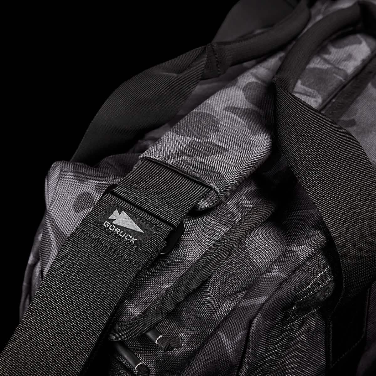 Close-up of the GORUCK Kit Bag, featuring a black and gray camouflage pattern reminiscent of a Special Forces Flight Bag. It has sturdy black straps, includes a robust buckle, and bears the GORUCK logo tag, all set against a solid black background.