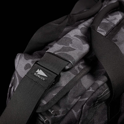 Close-up of the GORUCK Kit Bag, featuring a black and gray camouflage pattern reminiscent of a Special Forces Flight Bag. It has sturdy black straps, includes a robust buckle, and bears the GORUCK logo tag, all set against a solid black background.