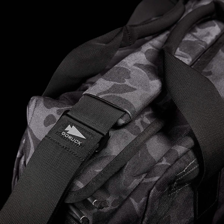 Close-up of the GORUCK Kit Bag, featuring a black and gray camouflage pattern reminiscent of a Special Forces Flight Bag. It has sturdy black straps, includes a robust buckle, and bears the GORUCK logo tag, all set against a solid black background.
