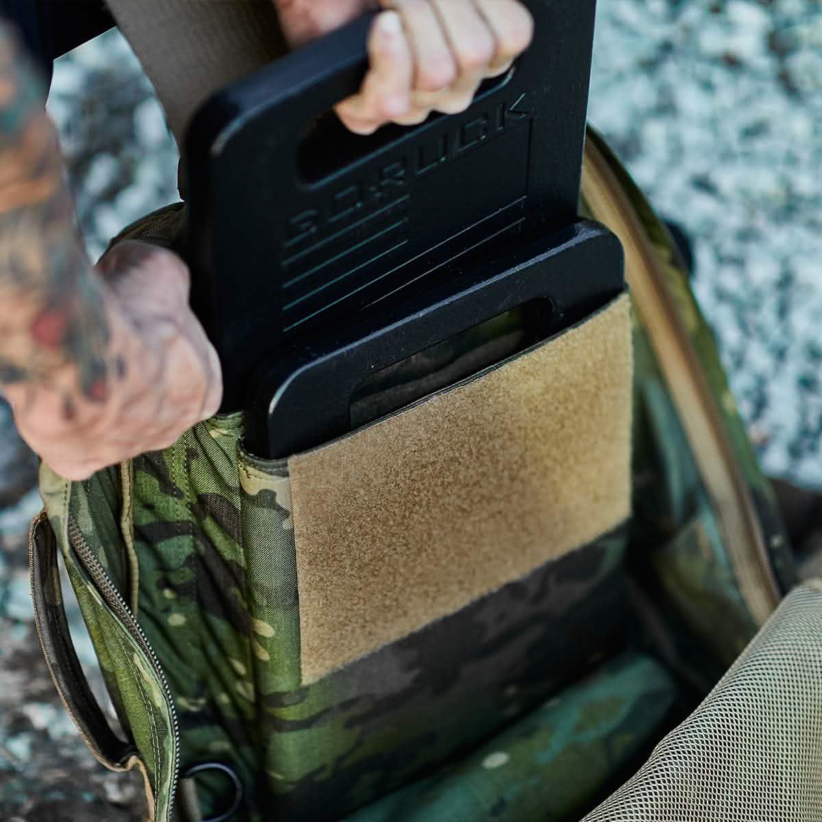 The tattooed arm slides a GORUCK Ruck Plate into the camouflaged backpack, using its Velcro divider to maintain organization. The scene, set against a textured stone surface, captures the rugged spirit of American manufacturing.
