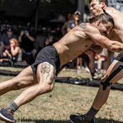 Two men wearing GORUCK's Men’s Ranger Panties - ToughStretch engage in an outdoor wrestling match on the grass. One, adorned with tattoos and wearing a knee brace, highlights the durability of ToughStretch fabric; the other sports short hair. Enthusiastic spectators look on eagerly, capturing moments with their cameras. The scene is lively and spirited, exemplifying a rugged essence backed by Scars Lifetime Guarantee confidence.