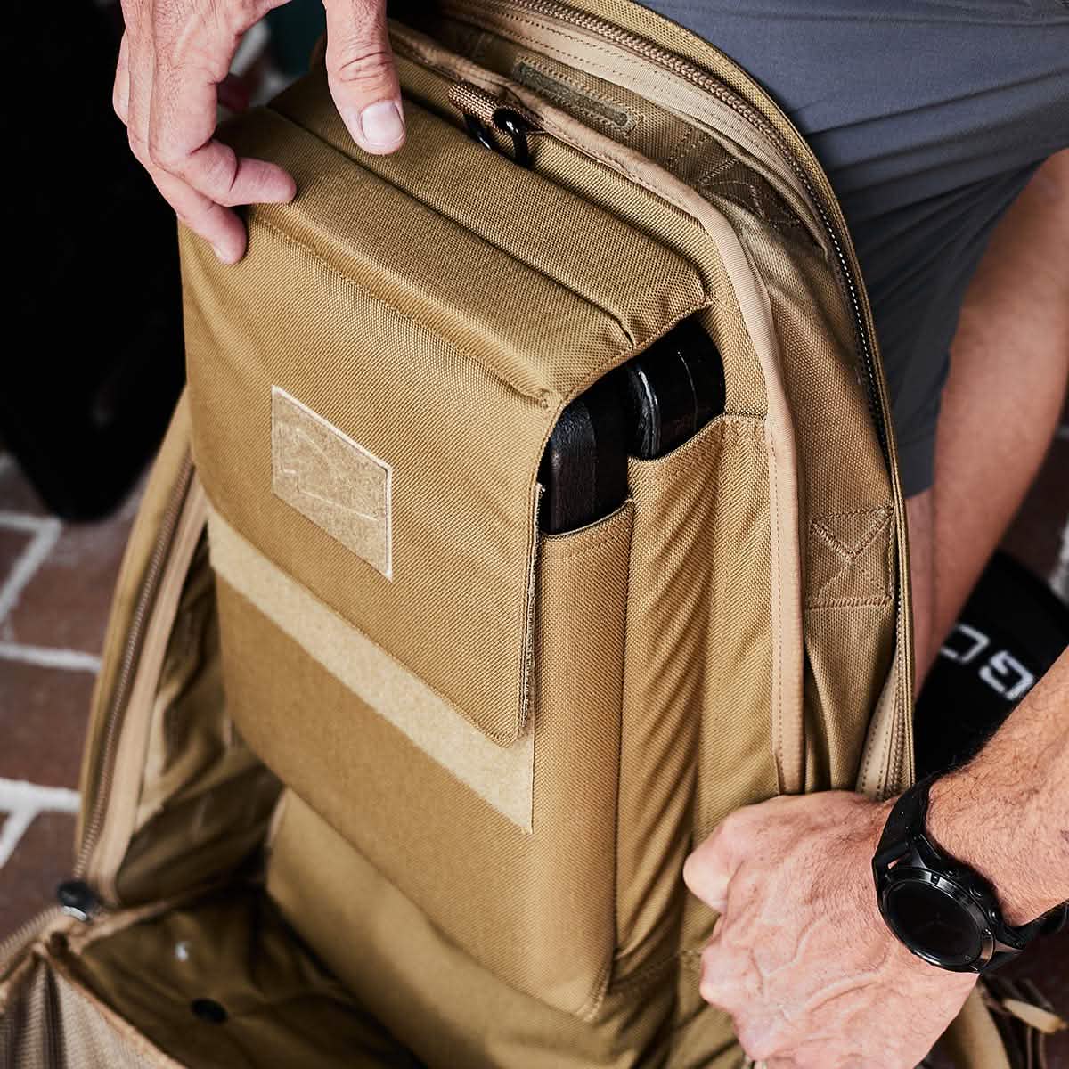 A person with a watch pulls out a padded compartment from a tan-colored GORUCK Rucker backpack. The compartment contains a black item, possibly a laptop. The bag rests on the brick surface as the person, wearing their gray shirt, showcases American manufacturing at its finest with their Ruck Plates.
