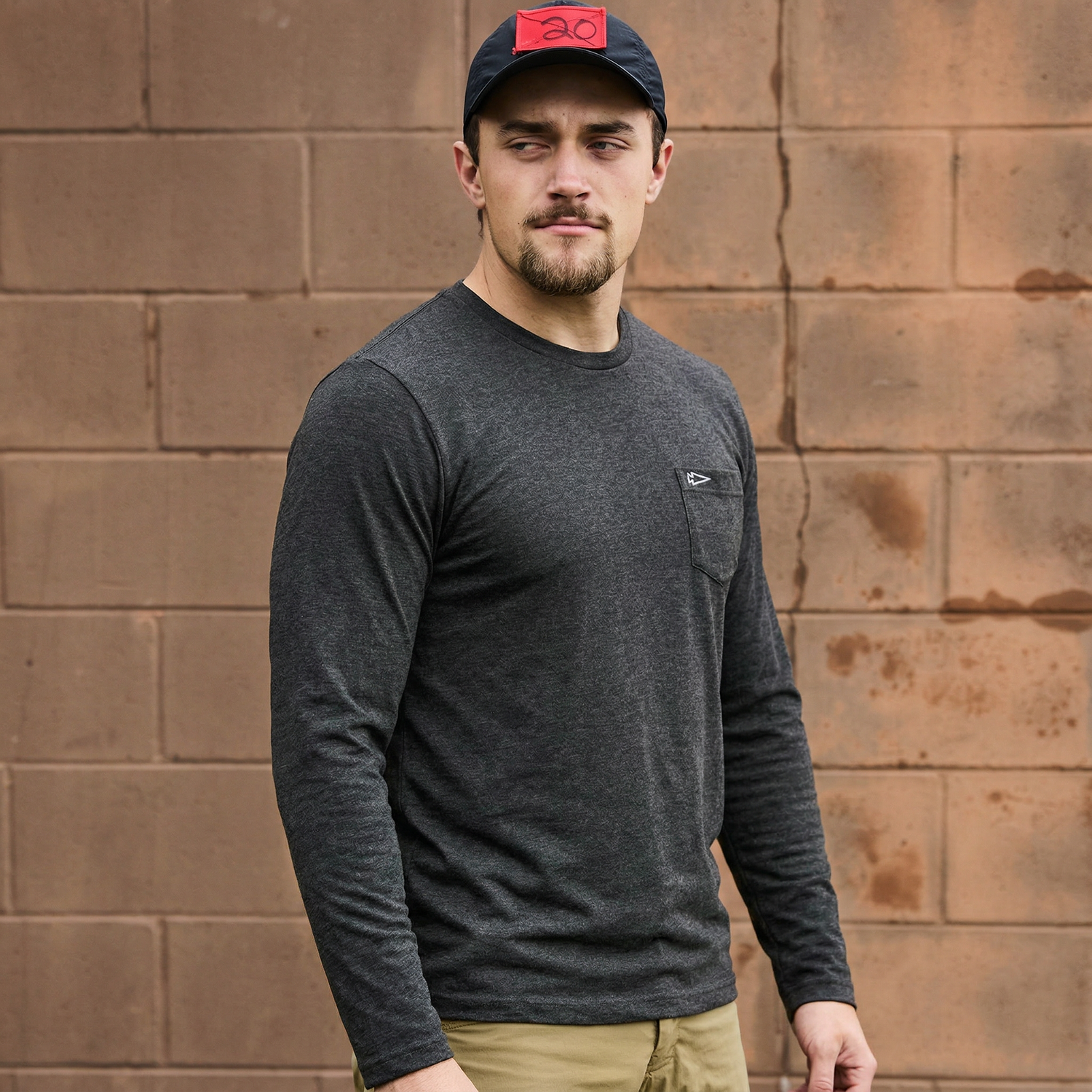 Man in a Men's Long Sleeve Pocket Tee - Tri-BlendX and black cap stands in front of a tan brick wall.