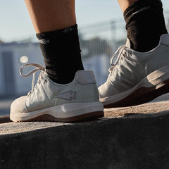A close-up shows someone wearing GORUCK's Men's Ballistic Trainers in Lunar Rock + Gum, featuring a silver reflective spearhead design. These athletic shoes are crafted from durable CORDURA Ballistic Nylon for exceptional wear, and the person is standing on a concrete ledge in black socks with a blurred fence in the background—ideal functional fitness footwear.